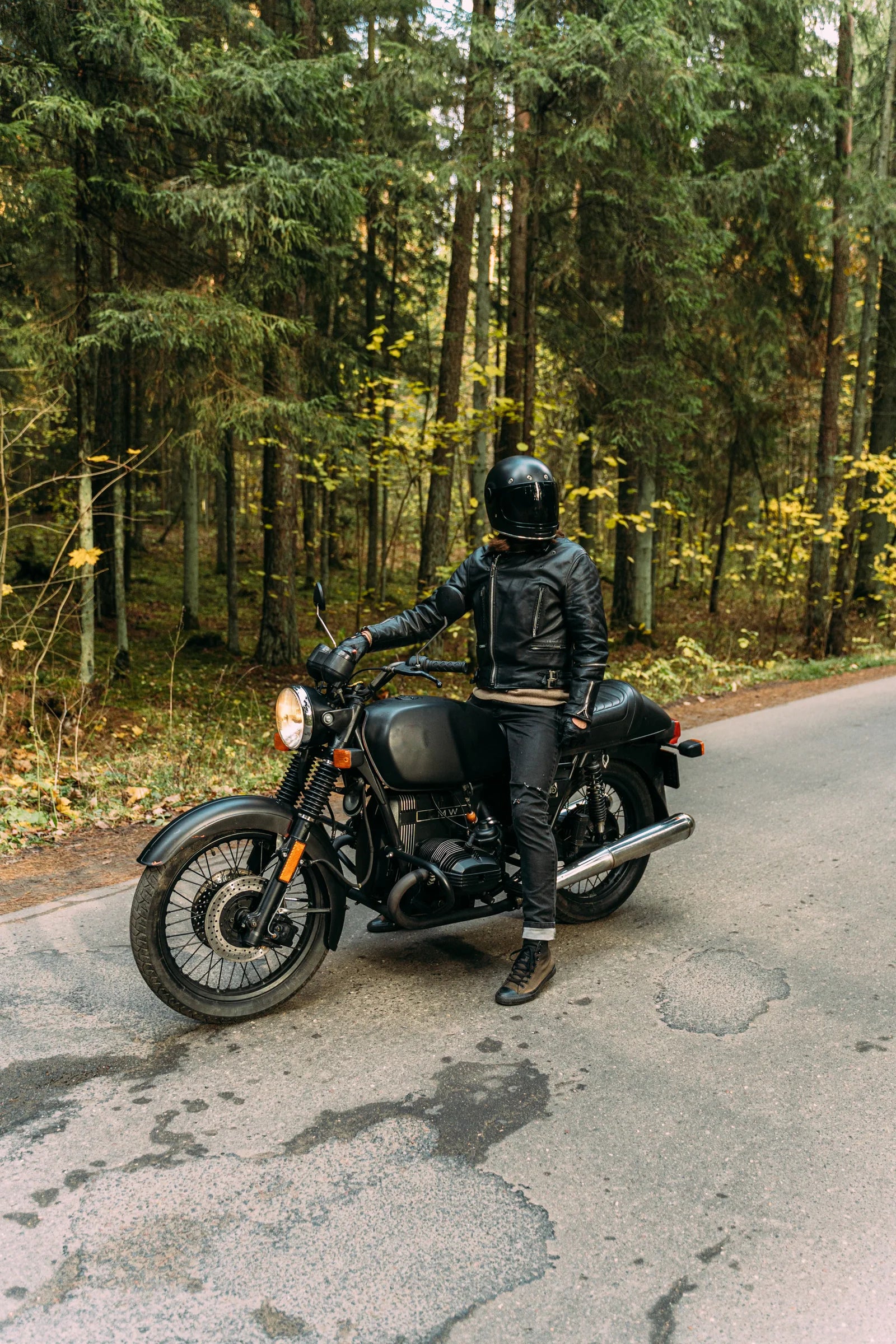 A helmeted rider with a black motorcycle beside a forest road.